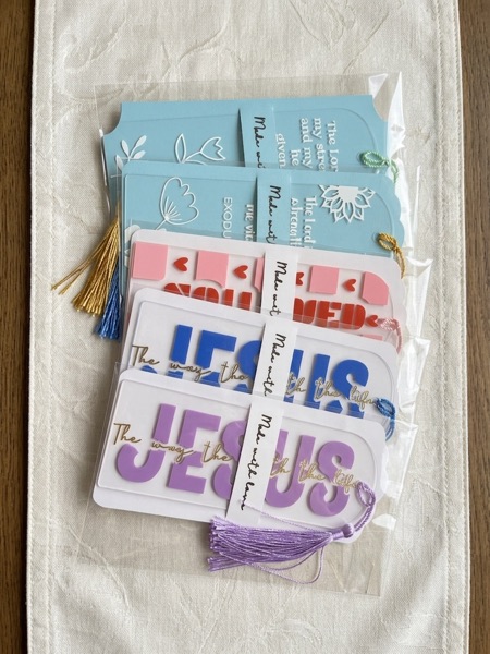 A fanned set of packaged bookmarks in blue, pink, white and lilac, including two lettered Jesus, each with a gold tassel.