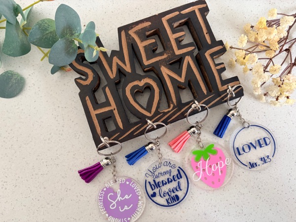 Round acrylic keychains with coloured tassels hanging from a Sweet Home sign, reading She is, Blessed and loved, Hope and Loved.