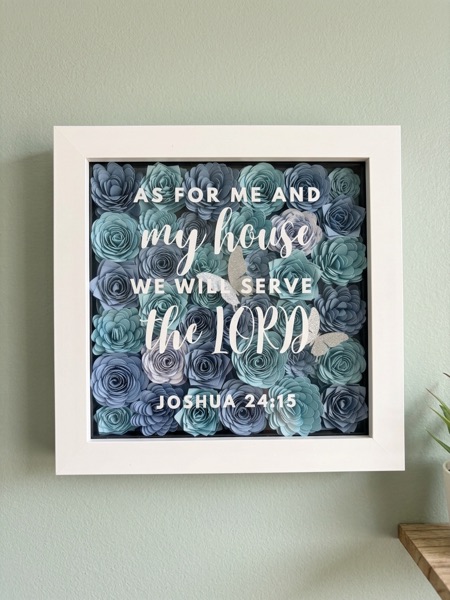 White shadow box filled with hand-rolled blue and grey paper flowers, lettered: As for me and my house, we will serve the Lord. Joshua 24:15.